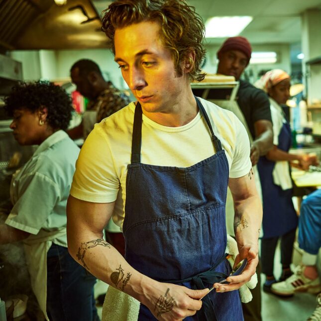 Inside a kitchen, Jeremy Allen White, who plays Carmy in the drama "The Bear," looks to the side while holding a spoon and dressing in an apron outside a white t-shirt. Working behind him from left to right are four other characters from "The Bear": Liza Colón-Zayas' Tina, Edwin Lee Gibson's Ebraheim, Lionel Boyce's Marcus, and Ayo Edebiri's Sydney. Ebon Moss-Bachrach's Richie sits on a kitchen counter, holding a cup on the right side of the frame — first opinion coverage from STAT