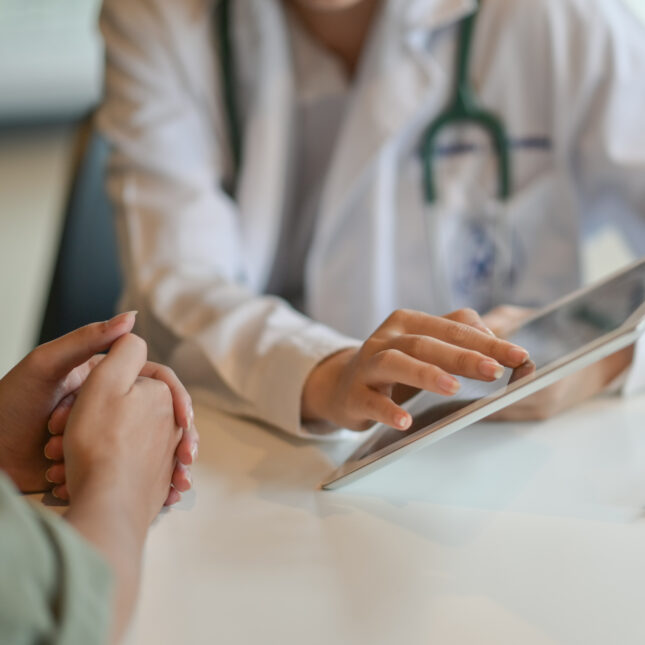 Photograph of a doctor wearing a white coat and stethoscope points to a tablet while speaking with a patient. -- health tech coverage from STAT