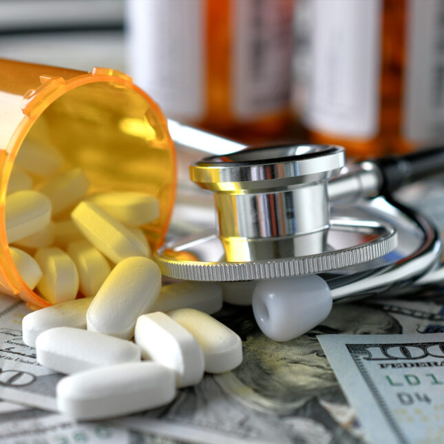 A stethoscope lays on several $100 bills, next to a laid-down pill bottle pouring out white oval pills and in front of two pill bottles standing up — first opinion coverage from STAT