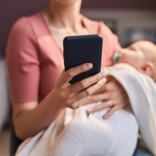 A person holds and looks at a smartphone while rocking a baby — coverage from STAT