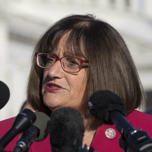 Photograph of Annie Kuster standing near mics and speaking to a crowd while wearing red glasses and a red blazer. -- health policy coverage from STAT