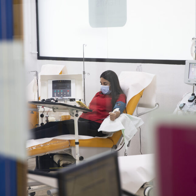 A masked person in red shirt and black pants sits in a donor bed, using their phone while donating blood plasma — first opinion coverage from STAT