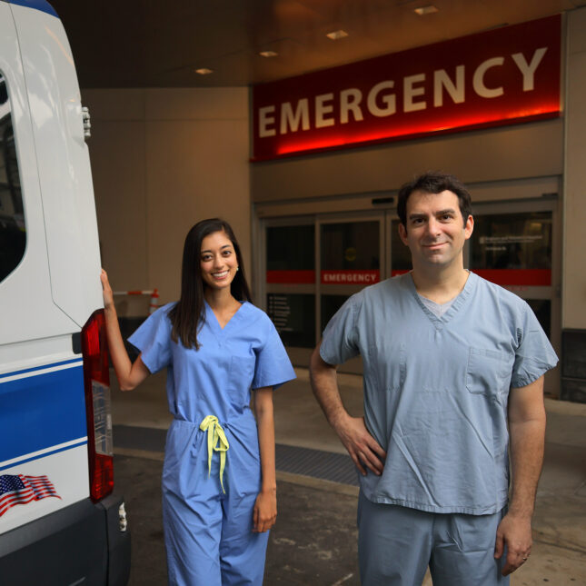 In front of an emergency room, a hospital resident stands in the middle with their hand touching the back of an ambulance. A teaching physician stands next to the new resident, putting a right hand on their own waist